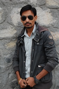 A person stands against a textured stone wall wearing sunglasses, a dark jacket, and a light-colored shirt. They have facial hair and are accessorized with a wristwatch and colorful bracelets.