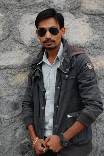 A person stands against a textured stone wall wearing sunglasses, a dark jacket, and a light-colored shirt. They have facial hair and are accessorized with a wristwatch and colorful bracelets.