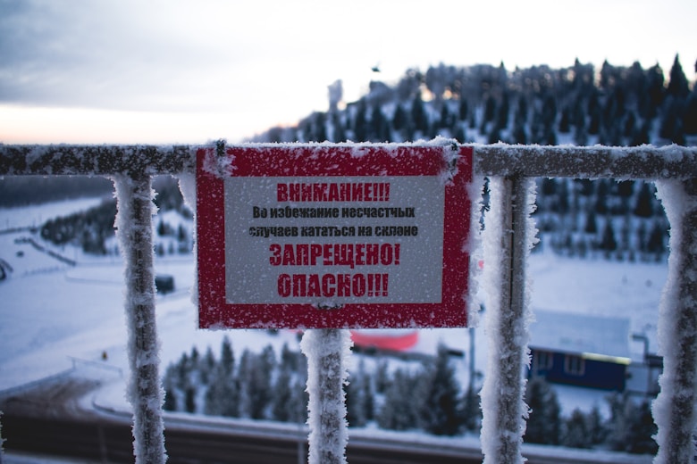 A warning sign with Russian text is attached to a frost-covered railing. The background displays a snow-covered landscape with trees and structures partially visible under a cold, overcast sky.