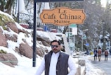 A man stands outdoors in a snowy environment near a sign that reads 'Le Cafe Chinar Murree.' He is wearing sunglasses and a dark jacket over a white garment. The background features snow-covered rocks, trees, and a few people walking on a snow-lined path.