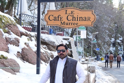 A man stands outdoors in a snowy environment near a sign that reads 'Le Cafe Chinar Murree.' He is wearing sunglasses and a dark jacket over a white garment. The background features snow-covered rocks, trees, and a few people walking on a snow-lined path.