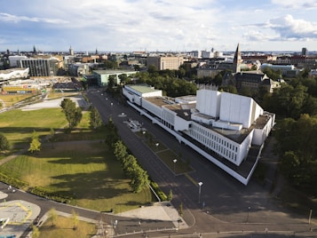 An aerial view of a cityscape featuring a prominent modern white building surrounded by trees and greenery. The structure has a geometric design with multiple tiers and sharp angles. In the background, a variety of buildings with different architectural styles can be seen, along with open parks and roads. The sky is partly cloudy and the sun casts long shadows, indicating late afternoon.