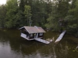 A wooden cabin is partially submerged in a calm, forested lake, connected by a narrow wooden bridge. The surrounding area is dense with trees and foliage, creating a secluded and rustic atmosphere.