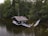 A wooden cabin is partially submerged in a calm, forested lake, connected by a narrow wooden bridge. The surrounding area is dense with trees and foliage, creating a secluded and rustic atmosphere.