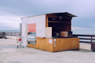 A small, rustic wooden kiosk with partly open sides, situated on a concrete waterfront overlooking the sea. The structure is simple, with a white tarp covering the back and a red element on one side. A solitary shopping cart is placed next to the kiosk, containing a red item. The atmosphere appears overcast, contributing to a subdued yet serene setting.