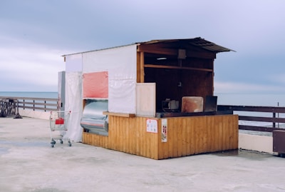 A small, rustic wooden kiosk with partly open sides, situated on a concrete waterfront overlooking the sea. The structure is simple, with a white tarp covering the back and a red element on one side. A solitary shopping cart is placed next to the kiosk, containing a red item. The atmosphere appears overcast, contributing to a subdued yet serene setting.