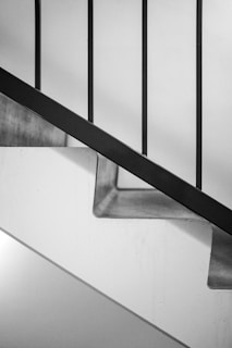 A modern staircase with a minimalist design, featuring a simple geometric shape and clean lines. The image is primarily in grayscale, highlighting the contrast between the dark metal railing and the lighter concrete steps.