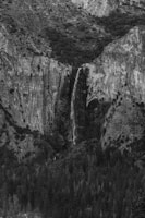 A dramatic black and white photograph captures a tall, narrow waterfall cascading down a rugged cliff face surrounded by dense forest. The textured rock formations and the dark foliage create a stark contrast, emphasizing the natural beauty and grandeur of the landscape.