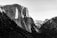 A dramatic black and white photograph of a mountainous landscape, featuring prominent cliffs and rugged terrain. The play of light and shadow highlights the textures of the rock surfaces.