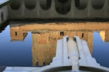 A water reflection of a building resembling a castle or fortress, captured in a calm body of water. The structure appears with arched openings and a mix of earthy, stone textures. In the foreground, there is a symmetrical stone feature partially submerged in the water, adding depth and leading the eye into the reflection.