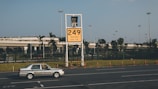 A car labeled 'Ground Service' is driving on a road in front of a large yellow sign displaying '249' with coordinates beneath. The background features a row of trees and a building with a rooftop garden, all under a clear blue sky.