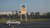 A car labeled 'Ground Service' is driving on a road in front of a large yellow sign displaying '249' with coordinates beneath. The background features a row of trees and a building with a rooftop garden, all under a clear blue sky.