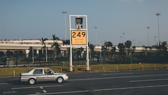 A car labeled 'Ground Service' is driving on a road in front of a large yellow sign displaying '249' with coordinates beneath. The background features a row of trees and a building with a rooftop garden, all under a clear blue sky.