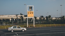 A car labeled 'Ground Service' is driving on a road in front of a large yellow sign displaying '249' with coordinates beneath. The background features a row of trees and a building with a rooftop garden, all under a clear blue sky.