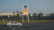 A car labeled 'Ground Service' is driving on a road in front of a large yellow sign displaying '249' with coordinates beneath. The background features a row of trees and a building with a rooftop garden, all under a clear blue sky.