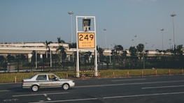 A car labeled 'Ground Service' is driving on a road in front of a large yellow sign displaying '249' with coordinates beneath. The background features a row of trees and a building with a rooftop garden, all under a clear blue sky.