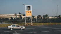 A car labeled 'Ground Service' is driving on a road in front of a large yellow sign displaying '249' with coordinates beneath. The background features a row of trees and a building with a rooftop garden, all under a clear blue sky.