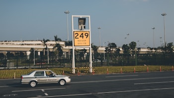 A car labeled 'Ground Service' is driving on a road in front of a large yellow sign displaying '249' with coordinates beneath. The background features a row of trees and a building with a rooftop garden, all under a clear blue sky.