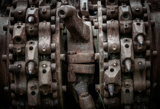 Complex machinery parts with heavy rust and wear, featuring interlocking gears and metal components. The image captures intricate details of industrial equipment showcasing bolts, nuts, and mechanical textures.