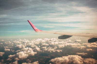 An airplane wing with a pink tip featuring a ribbon design is visible in the foreground. The wing cuts through a vast expanse of fluffy white clouds, under a soft sky that transitions from a light blue to a gentle beige hue.