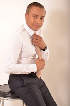 A man is sitting on a stool, dressed in a white shirt and dark pants, adjusting his tie. The setting appears neutral with a light-colored background.