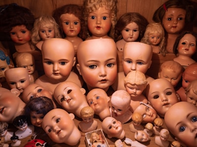 A collection of vintage doll heads displayed on a shelf, featuring various sizes and expressions. The dolls have diverse hair styles and colors, ranging from bald to curly blonde and brown wigs. Some dolls are older and more worn, contributing to an eerie atmosphere.