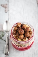 A jar filled with layers of red berry puree, oats, and topped with whole hazelnuts sits on a wooden surface. To the left, a metallic spoon is placed alongside the jar.
