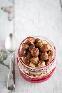 A jar filled with layers of red berry puree, oats, and topped with whole hazelnuts sits on a wooden surface. To the left, a metallic spoon is placed alongside the jar.