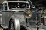 A vintage luxury car with a silver finish, featuring a prominent front grille and large round headlights. The vehicle showcases a classic, elegant design with intricate detailing.