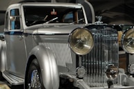 A vintage luxury car with a silver finish, featuring a prominent front grille and large round headlights. The vehicle showcases a classic, elegant design with intricate detailing.