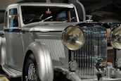 A vintage luxury car with a silver finish, featuring a prominent front grille and large round headlights. The vehicle showcases a classic, elegant design with intricate detailing.