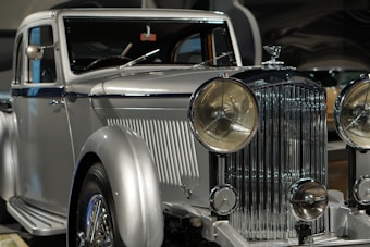 A vintage luxury car with a silver finish, featuring a prominent front grille and large round headlights. The vehicle showcases a classic, elegant design with intricate detailing.