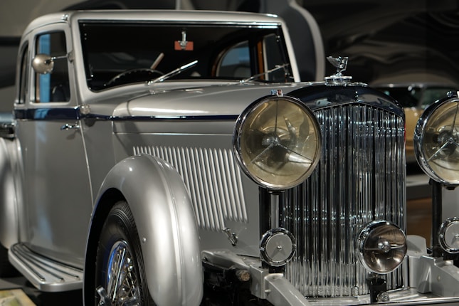 A vintage luxury car with a silver finish, featuring a prominent front grille and large round headlights. The vehicle showcases a classic, elegant design with intricate detailing.