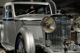 A vintage luxury car with a silver finish, featuring a prominent front grille and large round headlights. The vehicle showcases a classic, elegant design with intricate detailing.