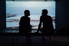Um homem e uma mulher sentados em um banco de uma sala escura e um telão ao fundo com imagem de natureza
