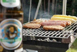 A variety of foods including sausages, burger patties, and corn on the cob are being grilled on an outdoor barbeque. In the foreground, there is a blurred image of a beer bottle. Smoke is rising from the grill, indicating the food is being cooked.