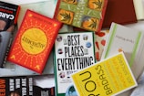 A collection of books is arranged haphazardly on a flat surface. The books are of various colors and titles, including 'The Alchemist,' 'The Best Places for Everything,' and 'You Are a Badass.' Some books are stacked on top of others, showing a mix of orientations with some covers facing up and others showing their spines.