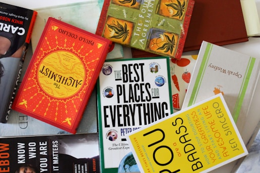 A collection of books is arranged haphazardly on a flat surface. The books are of various colors and titles, including 'The Alchemist,' 'The Best Places for Everything,' and 'You Are a Badass.' Some books are stacked on top of others, showing a mix of orientations with some covers facing up and others showing their spines.