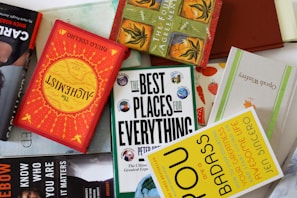 A collection of books is arranged haphazardly on a flat surface. The books are of various colors and titles, including 'The Alchemist,' 'The Best Places for Everything,' and 'You Are a Badass.' Some books are stacked on top of others, showing a mix of orientations with some covers facing up and others showing their spines.