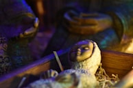 A peaceful nativity scene featuring a carved figure of a baby in a wooden manger surrounded by straw. Two figures with clasped hands are visible in the background, creating a sense of care and protection.