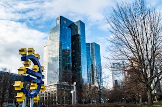 A collection of modern high-rise buildings with reflective glass exteriors stands prominently in the background. In the foreground, a large blue euro currency symbol adorned with yellow stars is displayed, positioned in a public space with bare trees and cloudy skies.