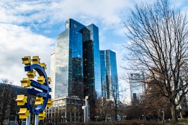 A collection of modern high-rise buildings with reflective glass exteriors stands prominently in the background. In the foreground, a large blue euro currency symbol adorned with yellow stars is displayed, positioned in a public space with bare trees and cloudy skies.