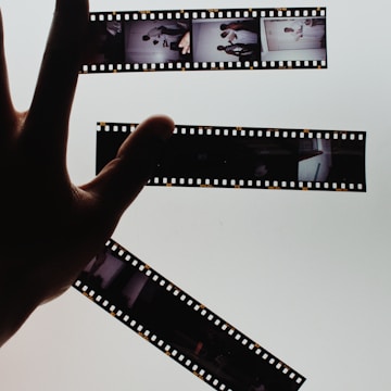 A hand is holding up strips of developed film negatives against a bright, light background, creating a strong contrast. The film strips contain several small images depicting various scenes.