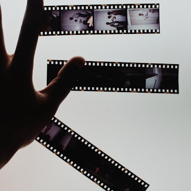 A hand is holding up strips of developed film negatives against a bright, light background, creating a strong contrast. The film strips contain several small images depicting various scenes.