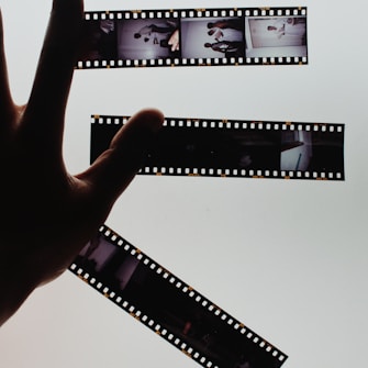 A hand is holding up strips of developed film negatives against a bright, light background, creating a strong contrast. The film strips contain several small images depicting various scenes.