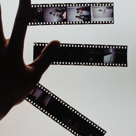 A hand is holding up strips of developed film negatives against a bright, light background, creating a strong contrast. The film strips contain several small images depicting various scenes.