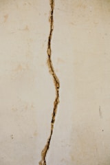 A vertical crack runs through the center of an off-white, textured wall. The crack appears irregular and jagged, exposing darker material beneath the surface.