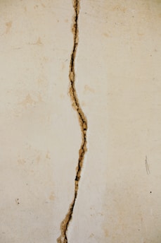 A vertical crack runs through the center of an off-white, textured wall. The crack appears irregular and jagged, exposing darker material beneath the surface.