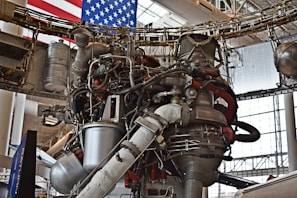 A complex assembly of machinery and pipes, possibly a rocket engine, is surrounded by various metal components and wiring. The background features part of an American flag, indicating a space or aerospace setting. Large, cylindrical objects and structural supports are visible alongside the intricate mechanical details.