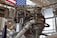A complex assembly of machinery and pipes, possibly a rocket engine, is surrounded by various metal components and wiring. The background features part of an American flag, indicating a space or aerospace setting. Large, cylindrical objects and structural supports are visible alongside the intricate mechanical details.